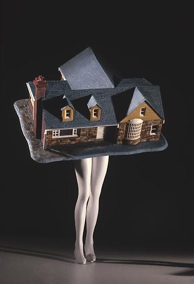 Foto di Laurie Simmons, raffigura una casa in minatura sorretta da un paio di gambe femminili bianche, simili a un manichino.