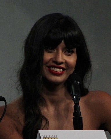 Jameela smiles with loose hair over bare shoulders, mic in foreground, in front of white background