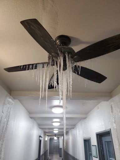 View looking down an indoor hallway. Icicles hang from a ceiling fan. Water or ice drips from the ceiling along the walls on both sides. The hallway is painted white, with blue along the lower part of the walls. A few blue doors line each side, and a few round light fixtures line the center of the hallway ceiling.