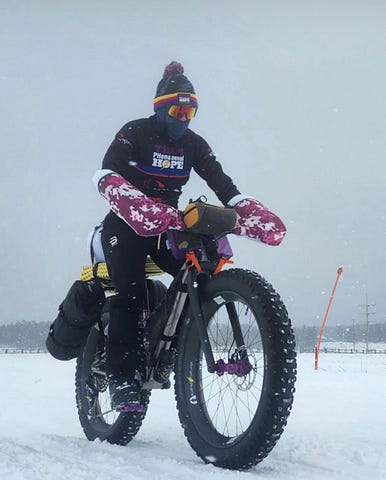 Patricia George, the article author, riding on a Why Cycles Big Iron fat tire bike in the snow with a Team PHenomenal Hope jacket and purple and pink camoflage pogies. The bike has all her camping gear on it as well.