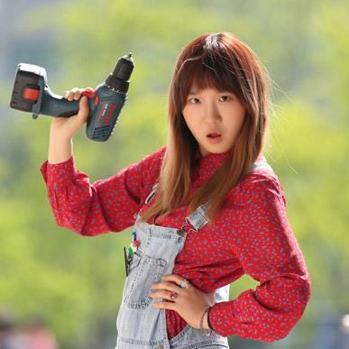 A woman in a red shirt and denim overalls holds up an electric drill.