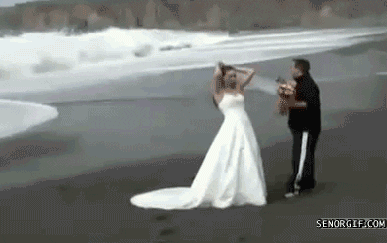 A bride and groom pose for a picture and a wave suddenly comes into the shore and knocks them both over.