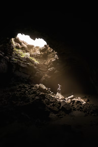 A person is in the well lit area from above of a cave, surrounded by rocks and blackness all around otherwise. They are looking down in spiritual pose.