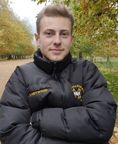 A photograph of Sam, wearing a black Oxford College puffer jacket, with his arms crossed. In the background is a tree-lined avenue.