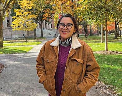 A young Diné woman wearing black-rimmed glasses, a purple sweater, brown jacket and black pants and boots standing on a sunny college campus with green grass and trees with green, yellow, and orange leaves