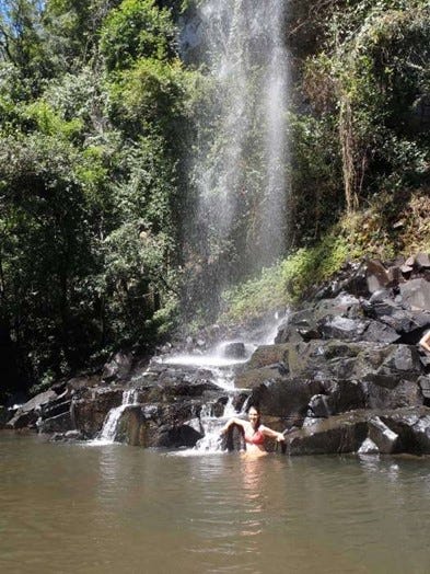 A girl swims in the base pool of a lightly spraying waterfall. There are a pile of rocks and the background is green rainforest.