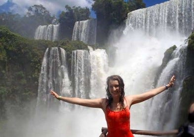 A girl in a pink tank top spreads her arms in front of several powerful waterfalls fringed with greenery