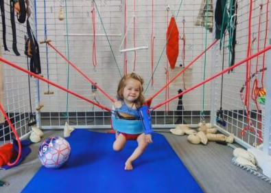 A girl is smiling while being supported by bungee cords to stand up independently during an intensive thearpy session while in a spider-cage unit.