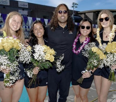 Photo of women’s swim team, adorned with flowers, smiling at the camera.