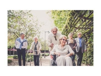 Three senior couples walking down a sunny street, one woman in a wheelchair.