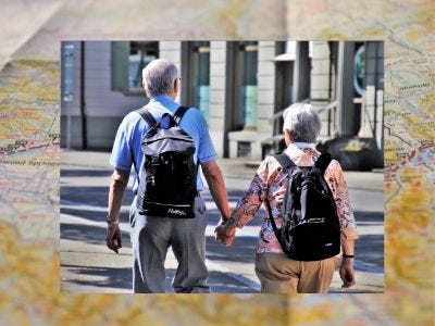 Rear view of a senior man and senior woman holding hands on vacation while wearing backpacks.
