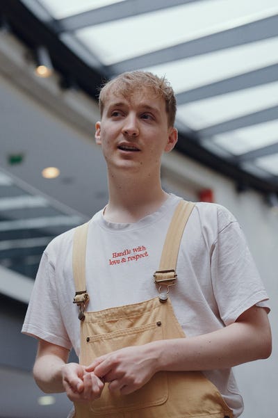Oli Isaac, a white nonbinary person with short blonde hair wearing a white shirt and yellow overalls. They look off to the side of the image and smile with their mouth slightly open as if in conversation.