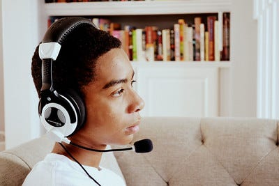 teen boy wearing headphones/microphone, staring to side with neutral face. short curly black hair, dark caramel complexion.