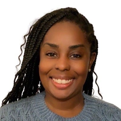 Photo of Berlin, a Black woman, from the shoulders up. She is looking at the camera with an open-lipped smile. She is wearing a blue sweater, with pink lipgloss and a stud earring visible on her right earlobe. Her long braids are pulled back behind her shoulders.