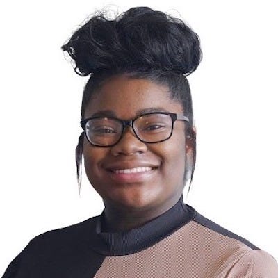 A photo of Mia, a black woman, from the shoulders up. She is looking at the camera with an open-lipped smile. She is wearing a black and brown top with her hair pulled into a high bun.