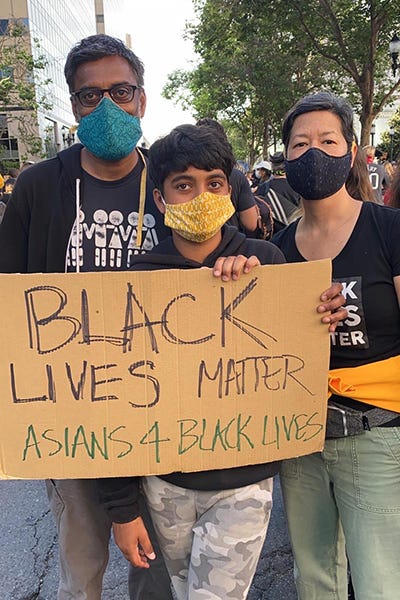 Two adults and a youth, all Asian, stand together at a rally with the sign: “Black Lives Matter #Asians4BlackLives.”