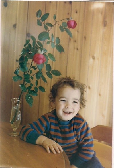 A photo of a smiling three year old child (Derek Neuland) with messy brown hair, wearing a red and blue striped shirt. Their arm is resting on a kitchen table. Next to them on the table is a tall clear glass vase with a flower in it, possibly a rose. Behind Derek is a wood paneled wall.