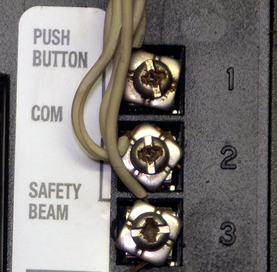 The back of a garage door motor is shown. Three screw  terminals are shown: “Push Button”, “COM”, “Safety Beam”.