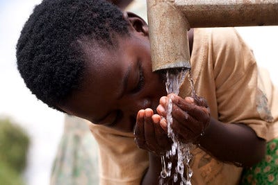 Young African boy cupping his hands to drink from a stream of water coming out of a pipe from a well