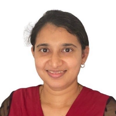 Photo of Supriya, a brown-eyed Asian woman, from the shoulders up. She is smiling at the camera. She is wearing a red top and her dark hair is pulled back with a side part.