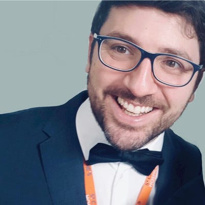 Peter is a white man, wearing blue glasses and a short beard. He has on a bowtie and a navy blue suit. He is smiling at the camera.