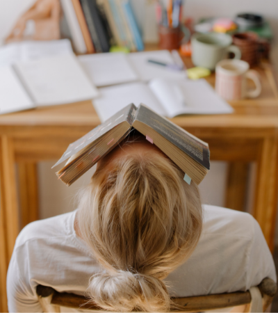 Overwhelmed student surrounded by books sleeping with book on head