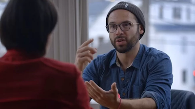 Photo of Aza Raskin being interviewed. He’s wearing glasses, facial hair, a beanie, and a button-down denim shirt as he gestures toward an interviewer at the edge of the frame.