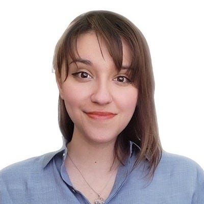 Photo of Alma, a Hispanic woman, from the shoulders up. She is facing directly into the camera with a closed-lip smile and curtain bangs framing her brown eyes. Her brown hair is straight and shoulder-length. She is wearing a collared, light blue button down shirt. She has winged eyeliner, peach lipstick, and a silver mushroom necklace.