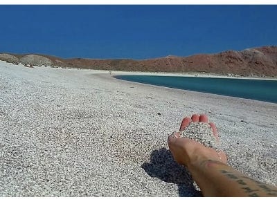 a photo of just a hand gathering sand on a beach, near turquoise water. the person appears to be laying down and the photo is taken from their perspective in Baja Mexico