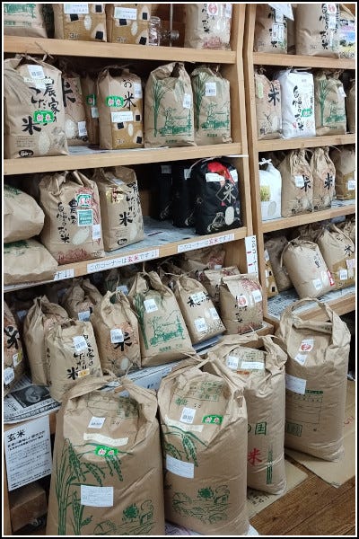 Grocery store shelves with brown 5kg bags of rice, each from a different farmer. The foreground shows 25kg bags of rice situated on the floor in front of the shelves.