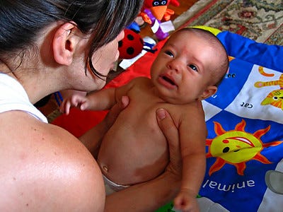 infant with a sad expression being held by woman with brown hair. baby wearing only diaper. colorful blanket in background.