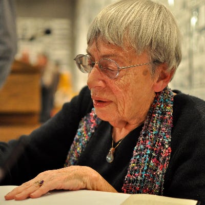 A photo of author Ursula K. Le Guin in her old age, wearing a black long sleeve shirt, multi-colored scarf, pearlescent necklace, and glasses. She has short cropped gray hair, and is in the midst of signing a book.