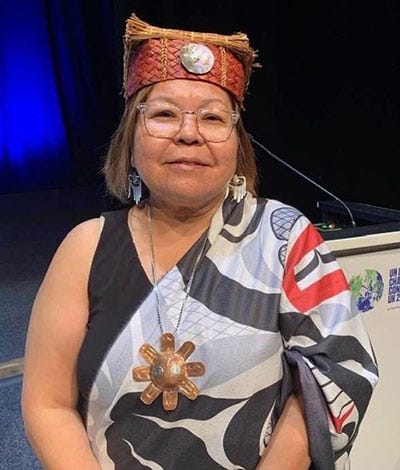 This is a photo of Leona Humchitt at COP26 in Glasgow, where she attended as a member of the Indigenous Clean Energy Advisory Board. She is using a red and brown hat, with a button on the front. A long, bronze-colored flower-shaped necklace and long white earrings that reach down to her neck. Behind her, we can see a podium.