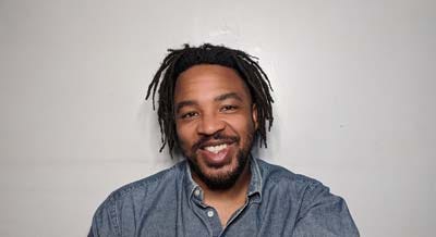 Bruce A. Lemon, Jr. wears a blue collared shirt and his hair in dreadlocks. He’s smiling directly at the camera for a portrait.