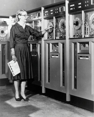 Black and white image of Grace Hopper examining in front of UNIVAC magnetic tape drives. She holds a COBOL programming manual in her hand.