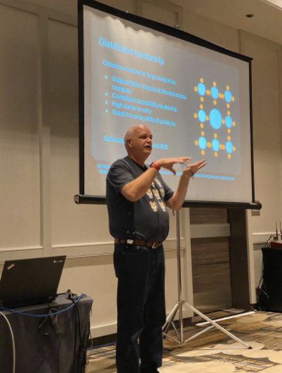 Ted Drake doing some hand gestures while presenting a slide in the backdrop saying distributed leadership with a diagram