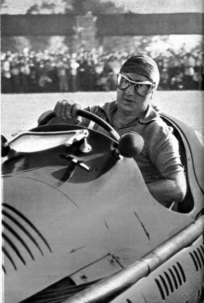 A black and white image of a slightly chubby guy driving a very old race car.