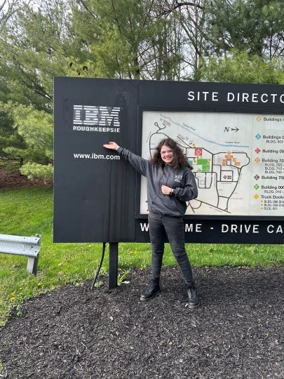 Cassie, dressed in a gray sweatshirt, black jeans, and black boots, smiles as she stands in front of and points to a large IBM Poughkeepsie site sign and map.