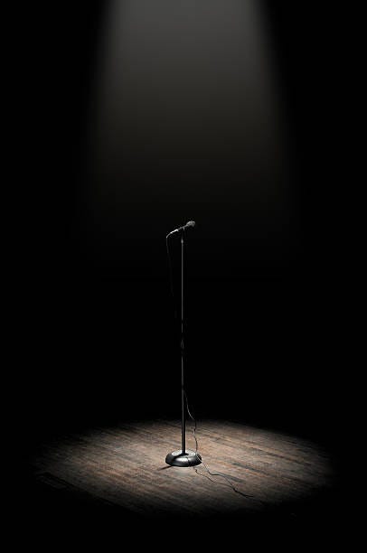 A mic on a microphone stand underneath a spotlight. Onstage. Background is all black except for the center stage, which is microphone stand on a brown, wooden stage.