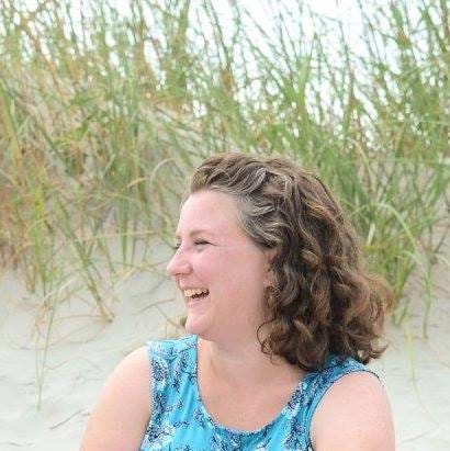 A woman with curly hair sits in the sand laughing. Behind her we see dune grass.