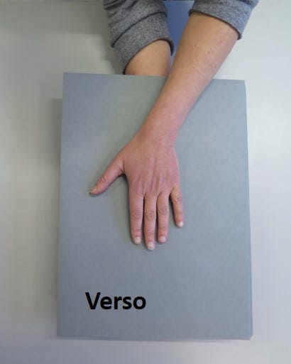 Two hands on either side of the same grey folder from the previous image. Here it has been flipped over to show the other side. The text in bottom left corner reads ‘Verso’