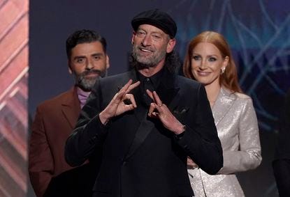 Troy is wearing a black suit and top hat, signing family. Behind him is Oscar Issac and Jessica Chastain, smiling against a blue background. Oscar is wearing a brown blazer and mauve turtleneck. Jessica is wearing a silver blazer and matching silver trousers.