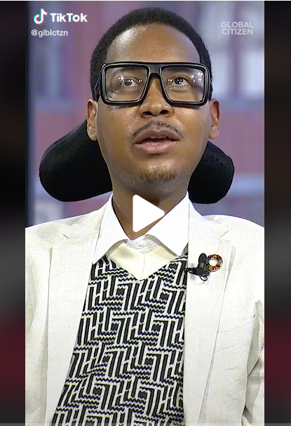 Still from a Tik Tok video of Black disability justice activist Eddie Ndopu. Eddie is seated in a wheelchair, wearing thick-framed black reading glasses, a white blazer, and sweater with a black and white geometric print. He is pictured mid-speech.