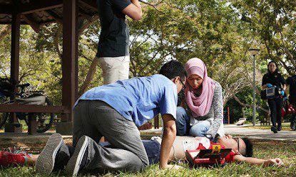 Man lying on the ground at a park while a woman and another man use an automated external defibrillator on him