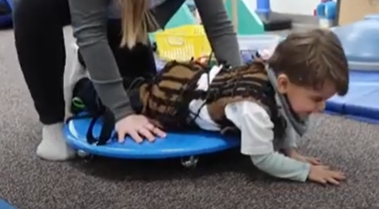 A therapist guides a child as he crawls while being supported by a moving thearpy scooter.