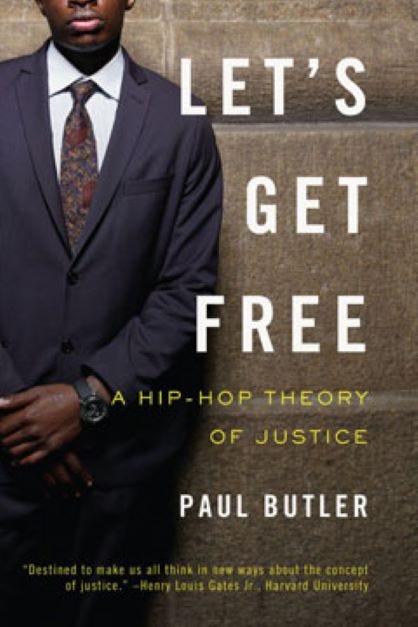 A book cover depicts a man in a dark suit and tie standing against a brick wall. Only the bottom half of his face is shown. White and yellow text reads, “Let’s Get Free: A Hip-Hop Theory of Justice, Paul Butler.”