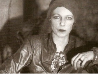 Photograph of Nancy Cunard, black-and-white, seated, wearing a leather jacket, a head-scarf and eyeliner.