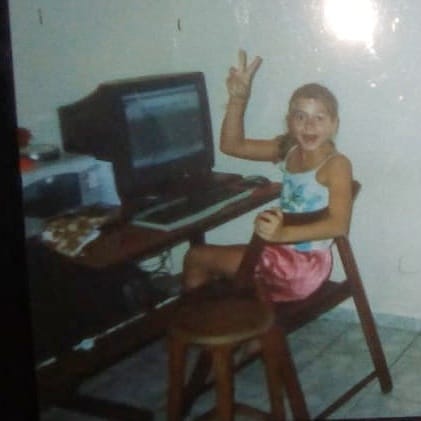 Garota sentada em cadeira usando o computador com monitor “de tubo” olhando para a camera sorrindo. Foto tirada de outra foto