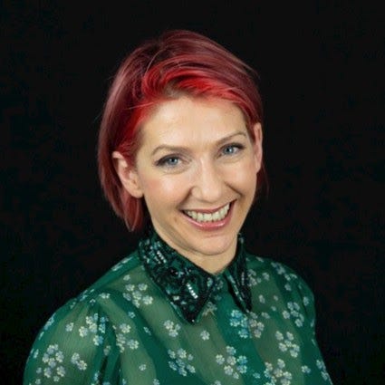 A white woman with pinky-red hair, wearing a green floral shirt and smiling at the camera.