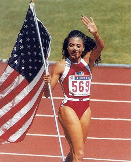 Flo Jo waves at the crowd while she bears the US flag in her red sprinting uniform.